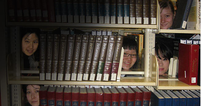 Students peaking out from behind books in the library stacks.